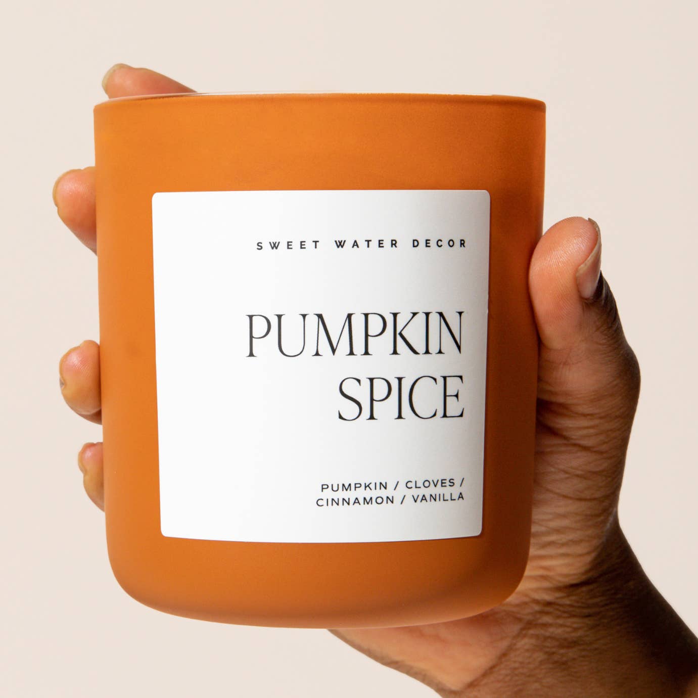 Orange candle labeled 'Pumpkin Spice' held by a hand against a beige background