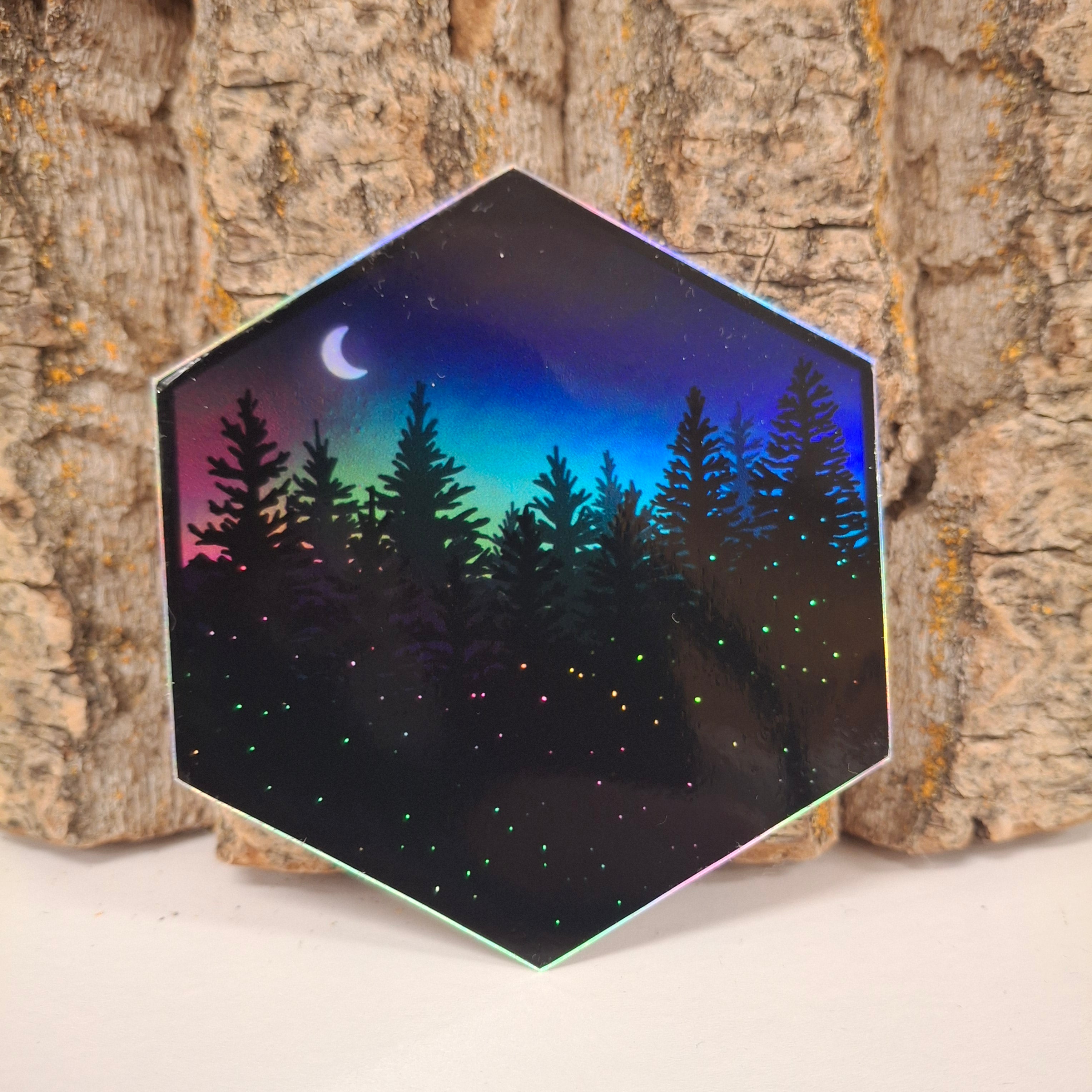 A hexagon sticker featuring the moon surrounded by twinkling stars and majestic pine trees. Holographic with a rainbow sheen. 