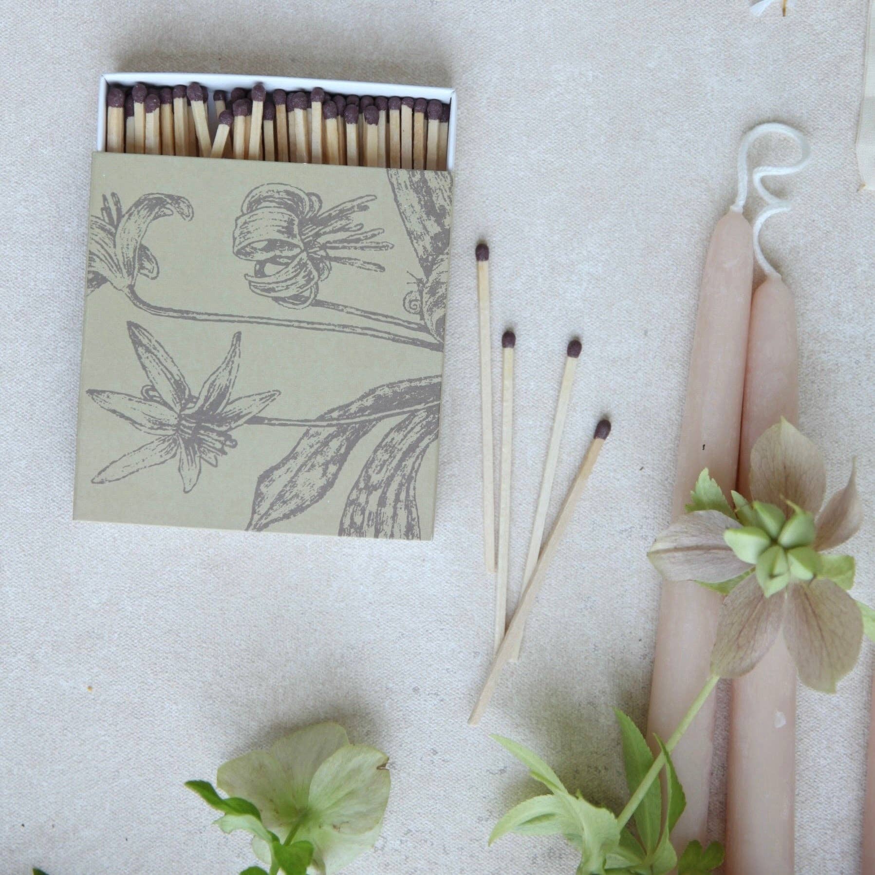 A matchbox with floral line art packaging sits on a light neutral surface beside three matches and two pale pink taper candles with green flowers arranged around them.