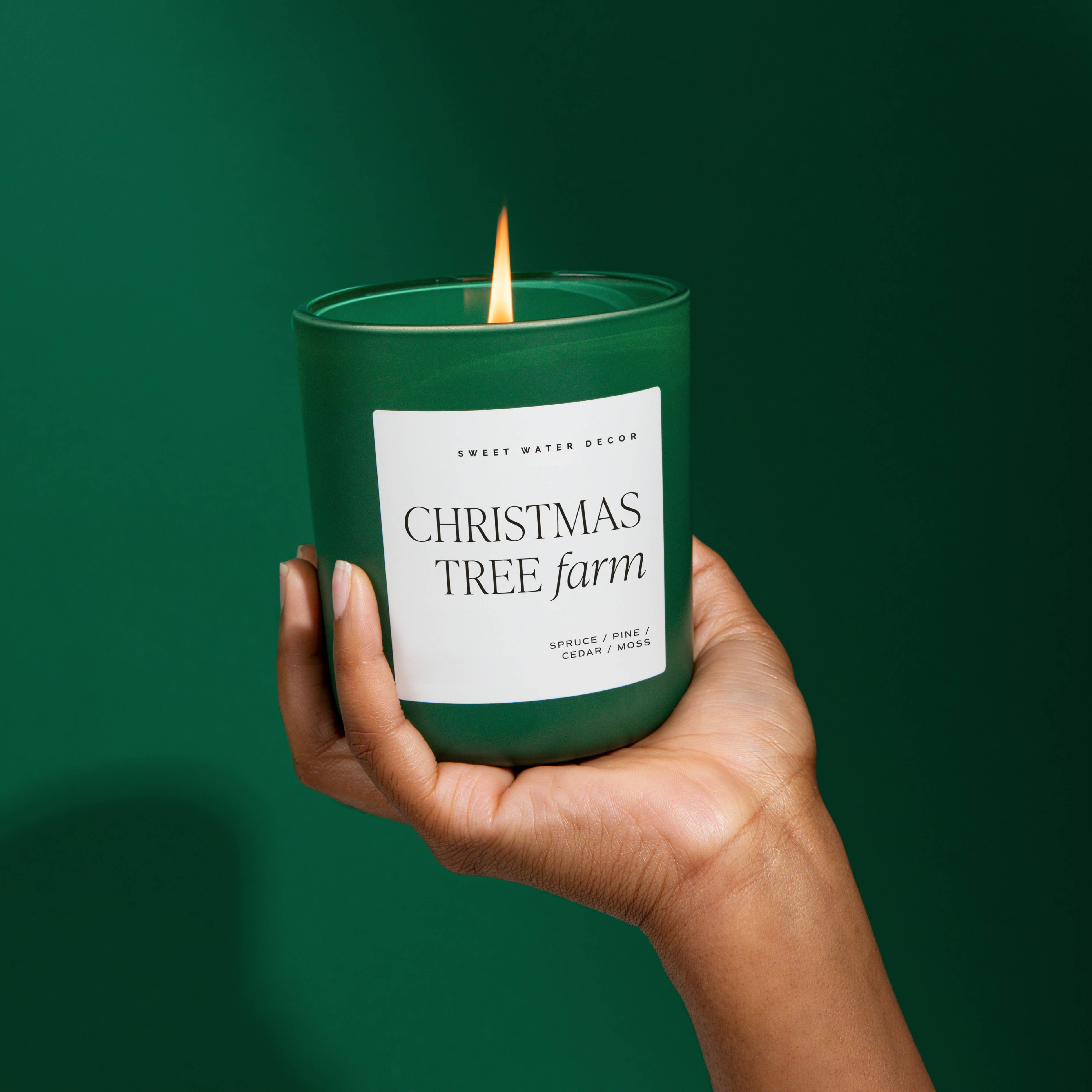 Hand holding a green candle labeled 'Christmas Tree Farm' against a dark green background