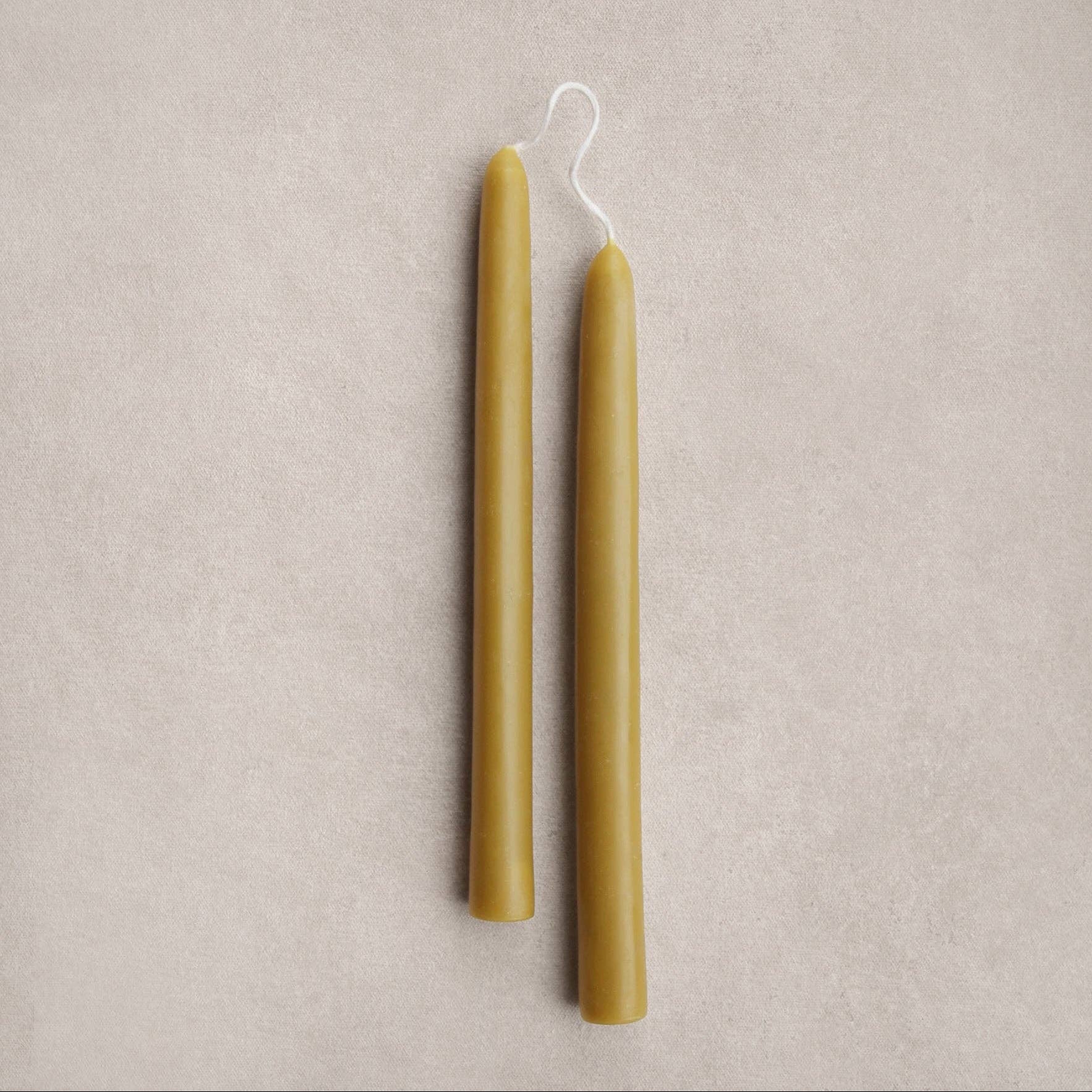 Two taper candles in a natural olive- wax, shown vertically on a textured light beige background.