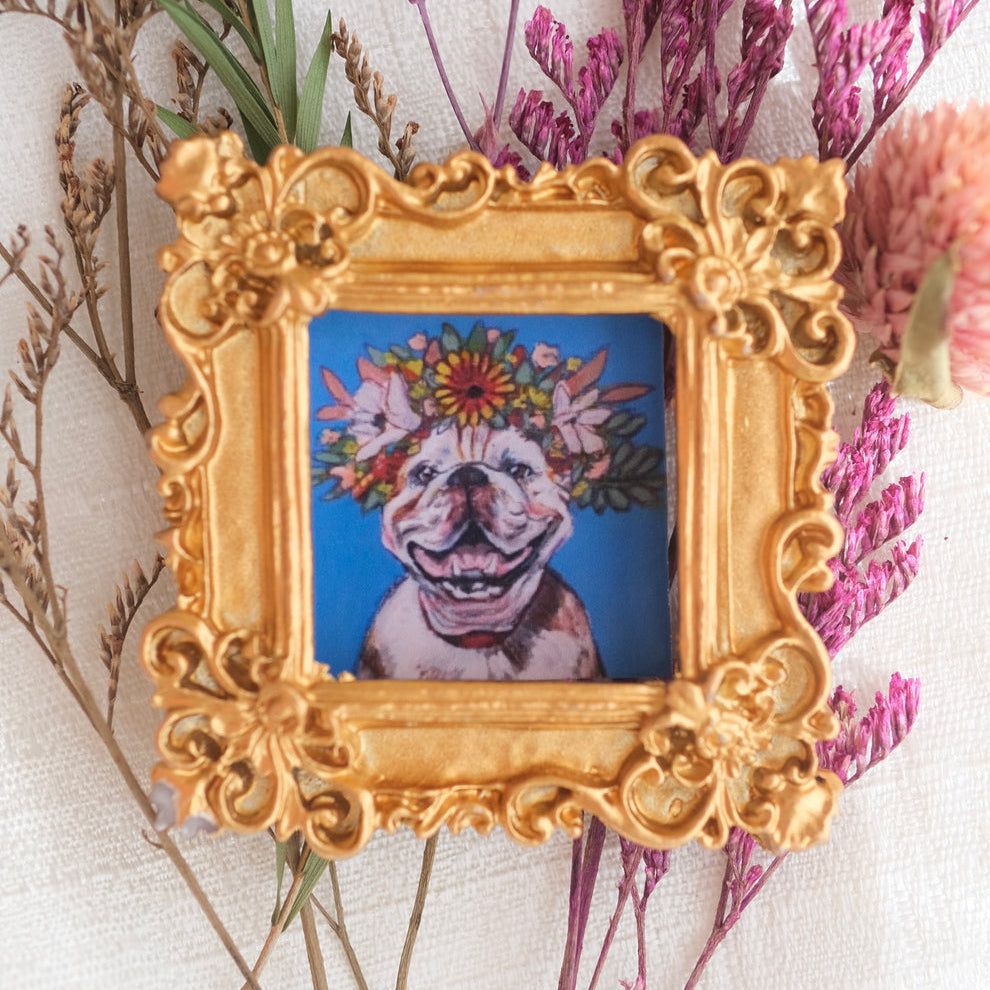 A small ornate gold frame features a cheerful painting of a smiling dog wearing a crown of colorful daisies and greenery. The artwork is set against a bright blue background and displayed with dried flowers around the frame for a whimsical, joyful presentation.