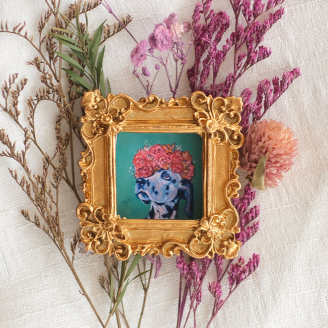 A small ornate gold frame displays a colorful painting of a dog wearing a crown of bright pink and red flowers. The frame is styled with intricate floral details and is surrounded by dried flowers on a textured cream fabric background.
