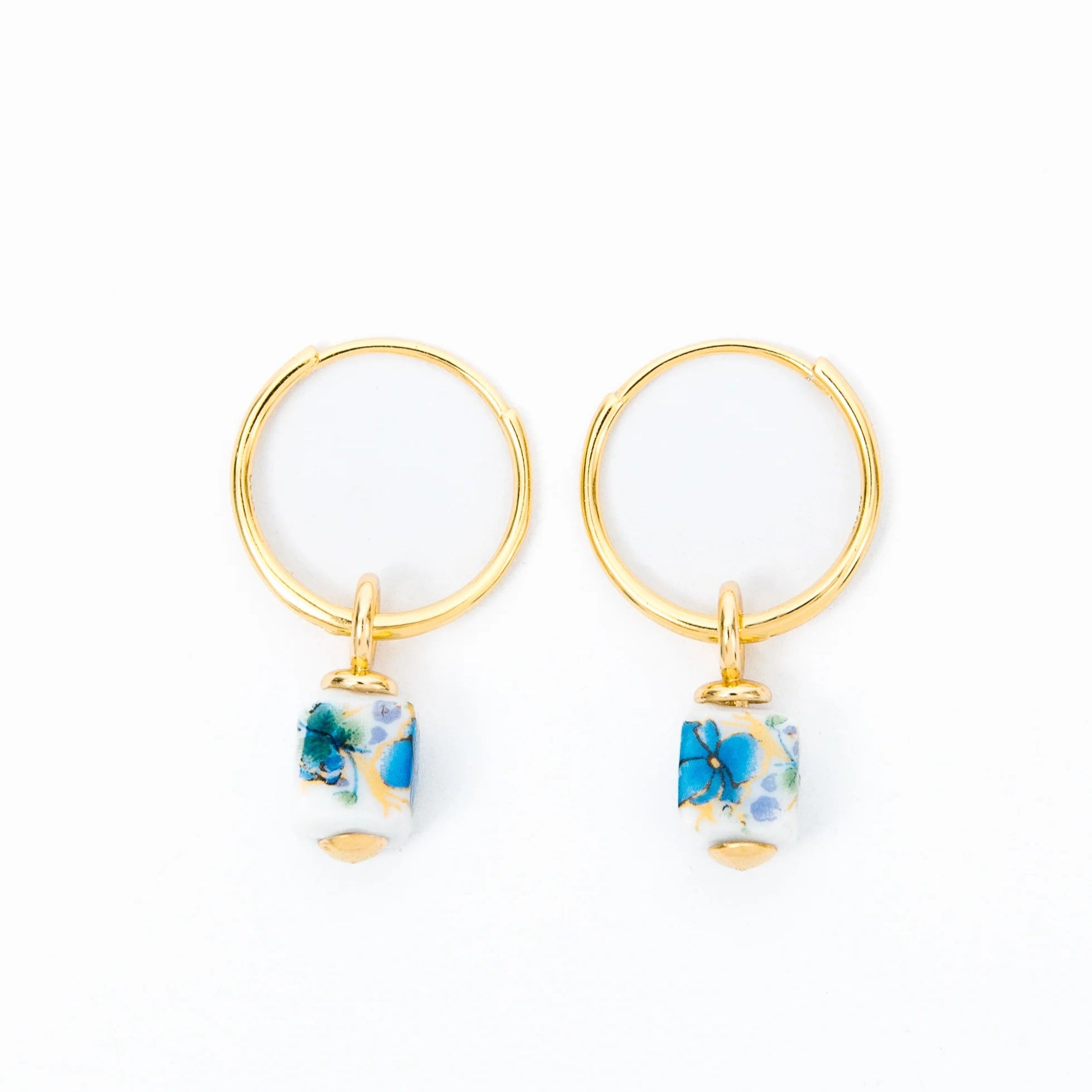 Gold hoop earrings featuring small, cylindrical ceramic charms with a delicate blue and green floral pattern. The colorful charms dangle from the hoops, adding a playful and elegant touch to the minimalist design.