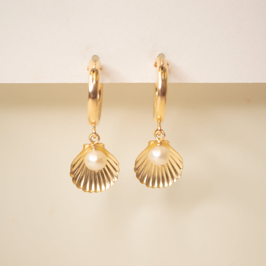 Gold hoop with a scallop sea shell and pearl dangle on an off white background.