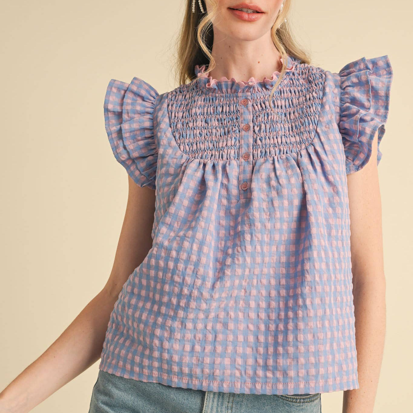 Embrace your softer side in this charming gingham flutter sleeve top. The delicate ruffled lace trim at the neckline and playful flutter sleeves create a romantic, feminine silhouette that feels both cheerful and flattering. A smocked yoke adds gentle structure, while the babydoll bodice offers comfortable, forgiving fit. The button-down placket makes it easy to style however you'd like—dress it up or keep it casual. Lined for confidence and comfort. Available in a lovely blue and pink.