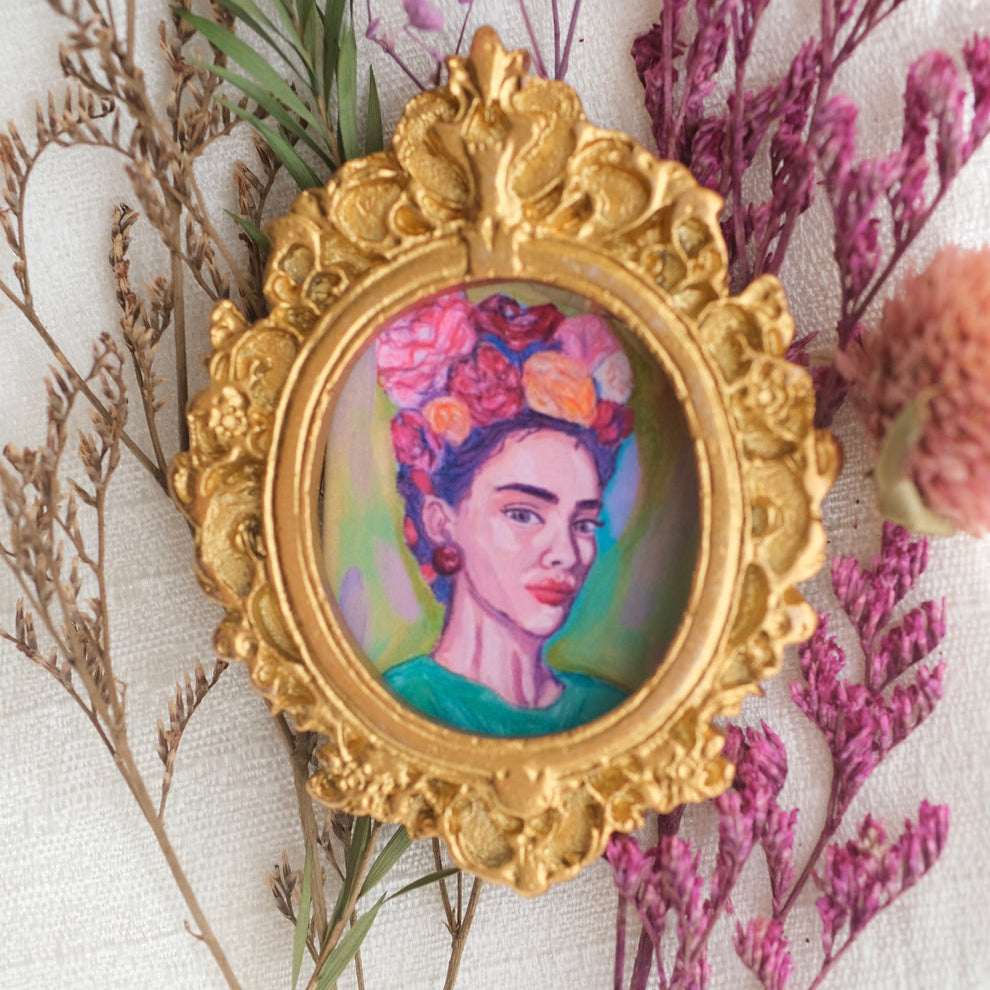 An ornate oval gold frame showcases a vibrant portrait of a woman wearing a crown of colorful flowers on her head. The artwork, placed on a textured fabric background with dried flowers, has a bold and artistic style that highlights her confident expression.
