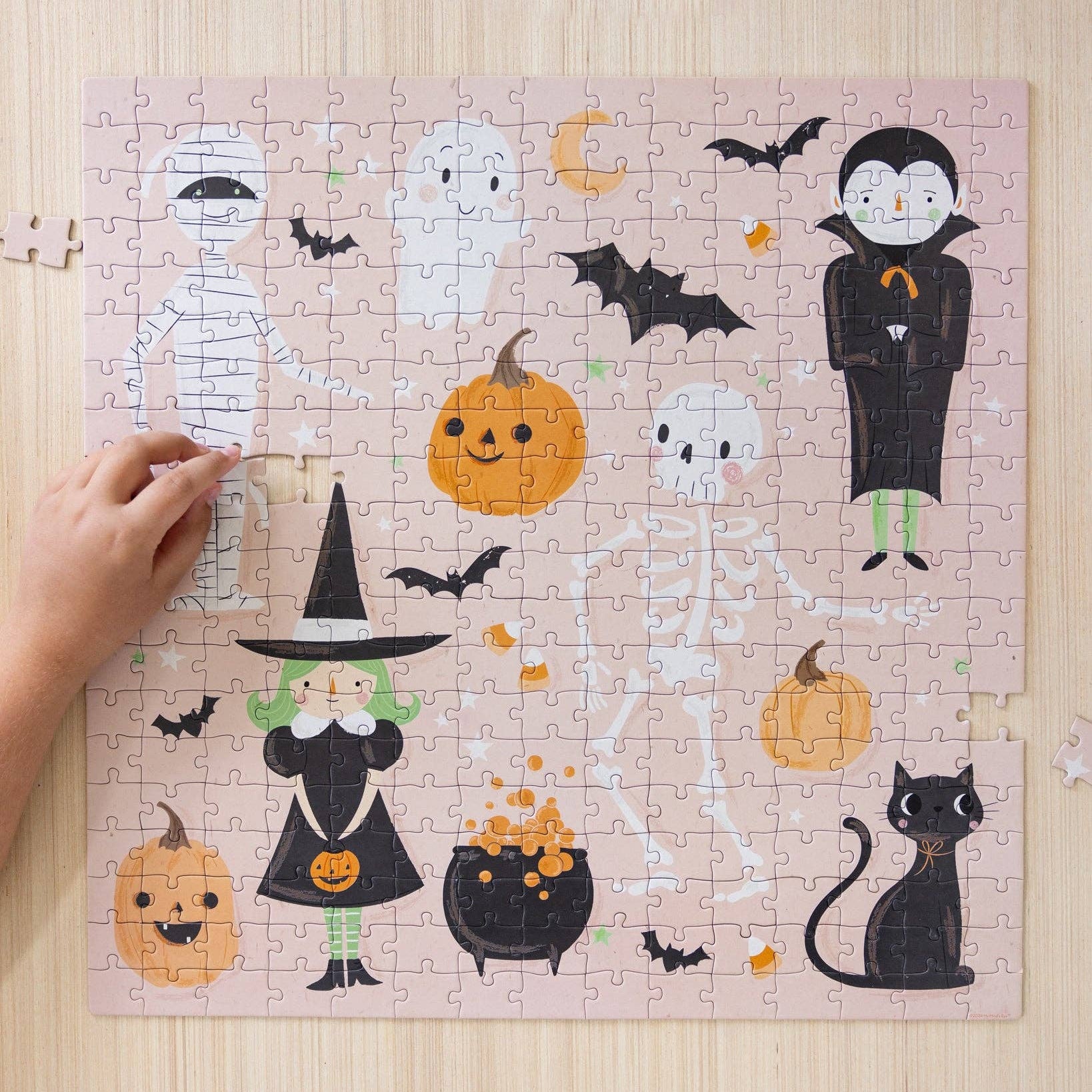 A completed rectangular jigsaw puzzle with a pastel pink background and Halloween-themed illustrations, including pumpkins, bats, a witch, skeleton, vampire, mummy, ghost, and cat. A person's hand is visible on the left holding a small piece, and the puzzle sits on a light wood surface.