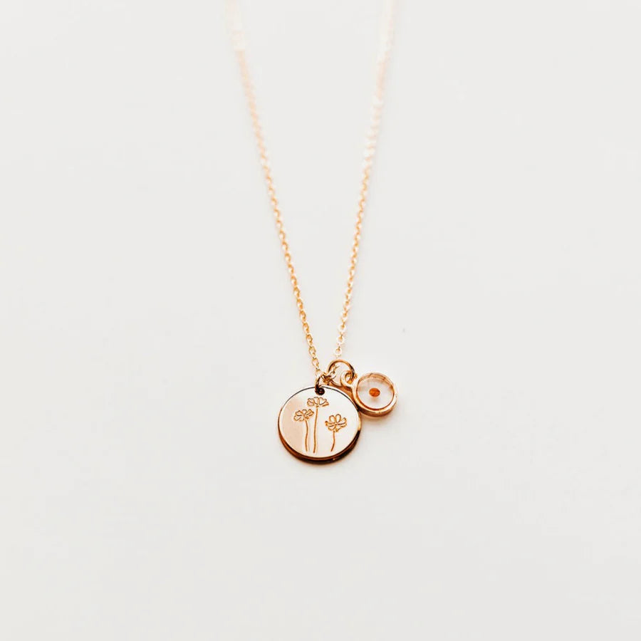 Gold necklace with round pendant featuring 3 individual buttercup flowers that have a hand drawn look to them. next to it is another round pendant featuring a mustard seen embezzled in a clear pendant