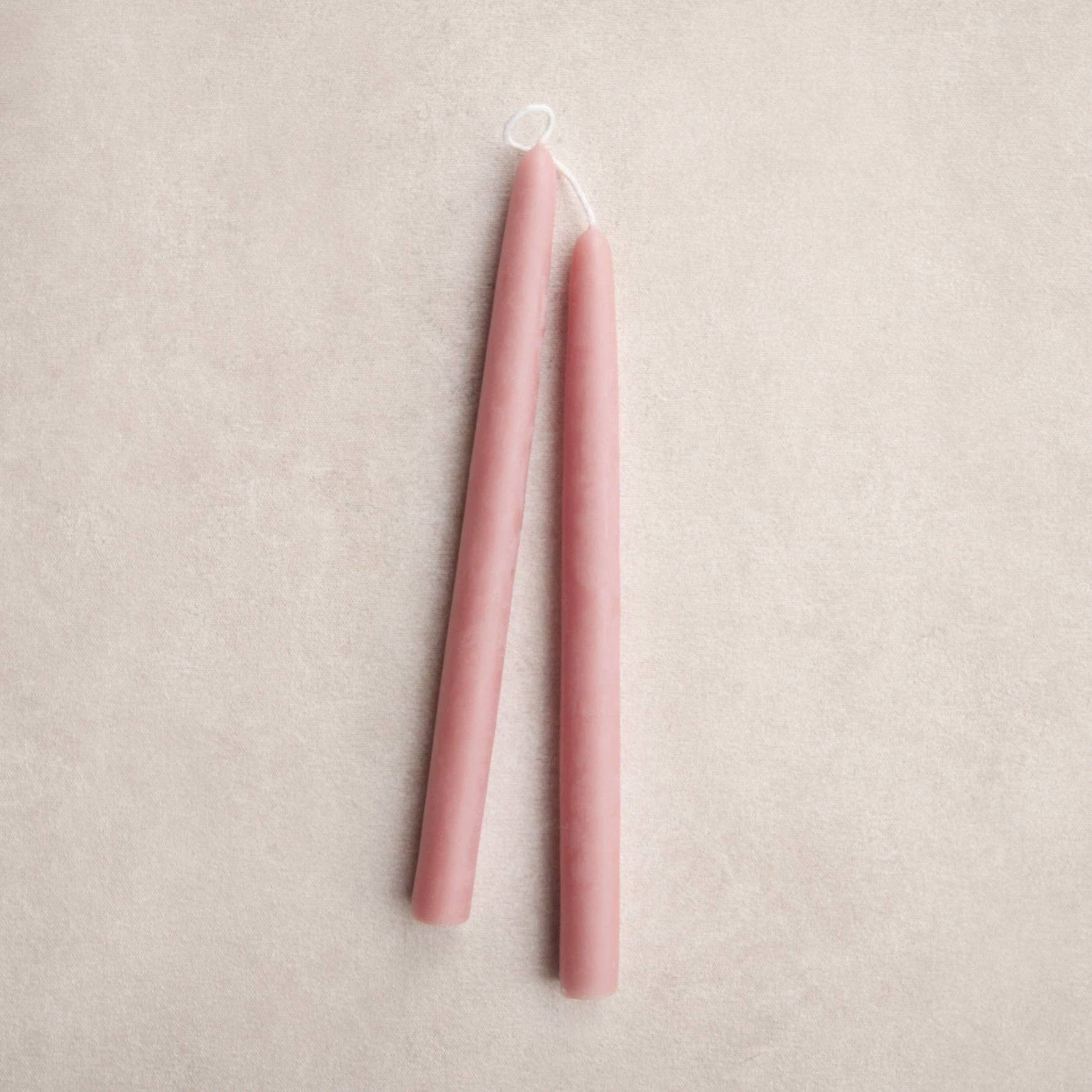 Two slim blossom color taper candles with white wicks, standing upright on a textured light beige background.