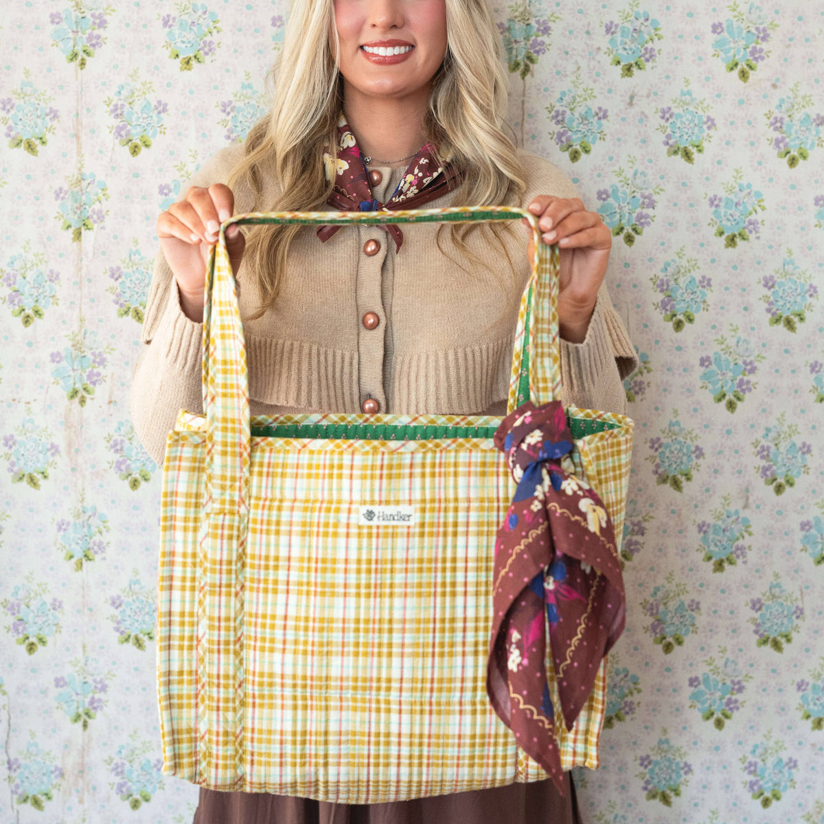 Woman holding a yellow plaid bag against a floral wallpaper background