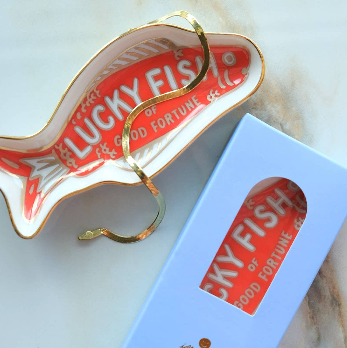 Ceramic fish-shaped trinket dish with gold rim and 'Lucky Fish' packaging on a marble surface.