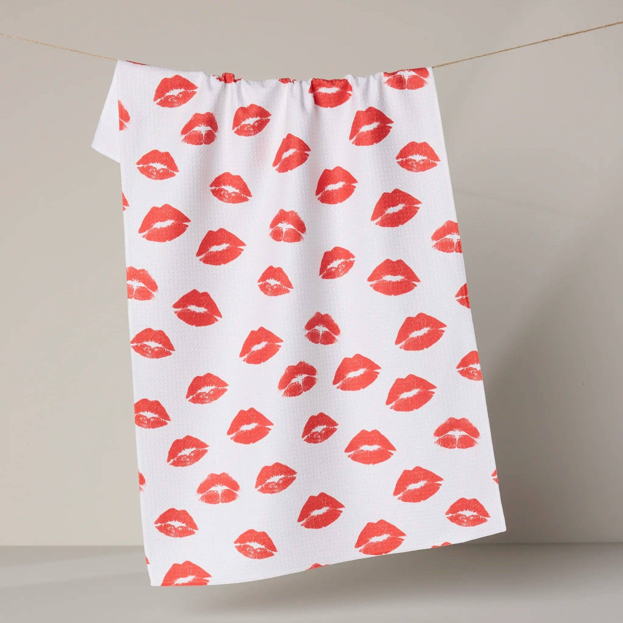 White towel with red lip print pattern hanging on a line against a neutral background