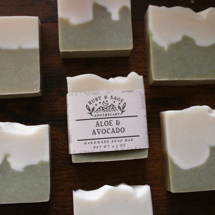A collection of handmade soap bars from Ruby & Sage Apothecary is arranged on a wooden surface, with one bar prominently labeled “Aloe & Avocado.” Each bar features a two-tone design with a soft green base and a creamy white top, reflecting the natural, nourishing ingredients.
