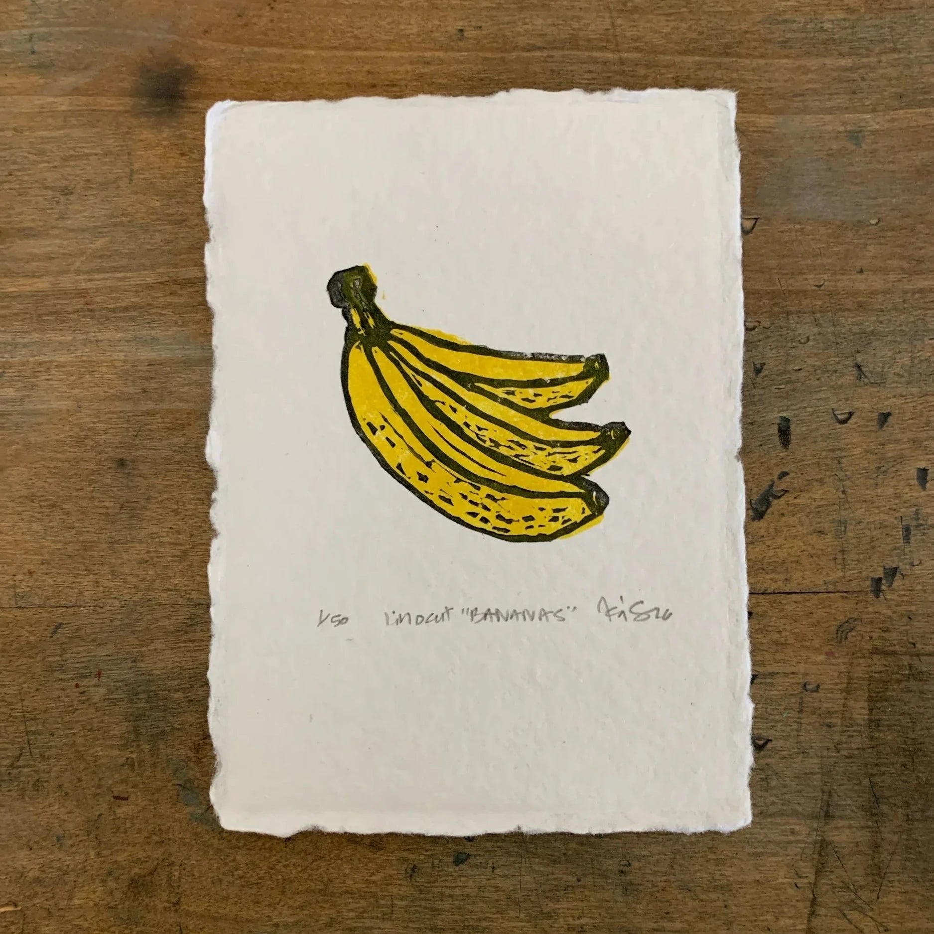 Yellow banana bunch on rough white paper. Signed by Kelsi, with title and date. Pictured against a wooden table. 