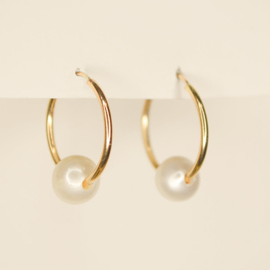 Thin gold hoops with a sliding white round pearl on each. Pictured on an off white background.