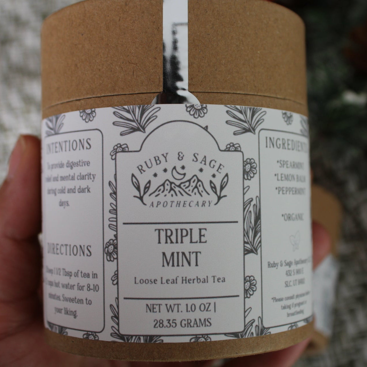 Brown container with a white label designed with sprigs and different fonts. Container held in a woman's hand, with a frosty green backdrop.