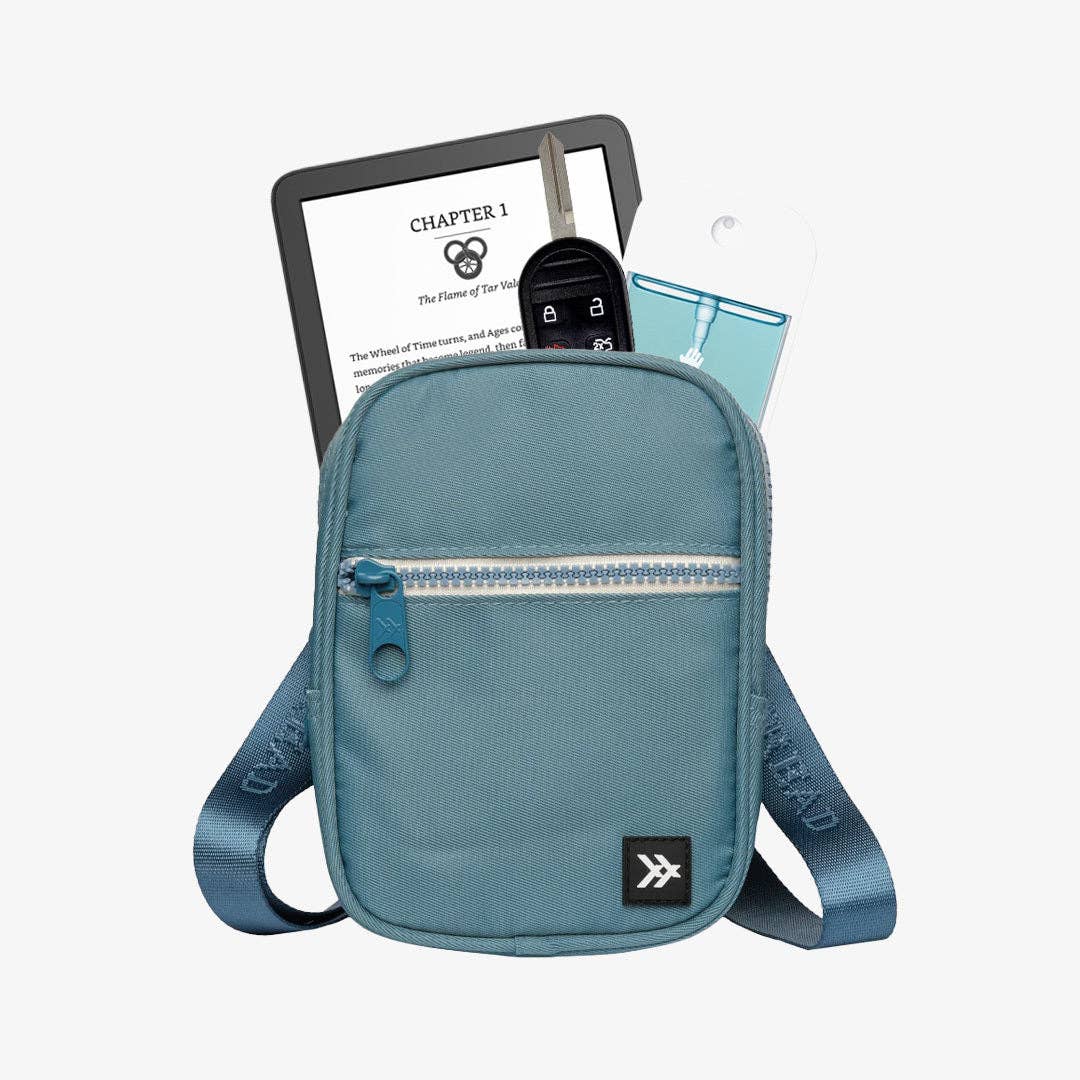 A light blue crossbody phone bag with a front zip pocket, shown with a tablet, keys, and a phone inserted into its side pocket, on a light gray background.