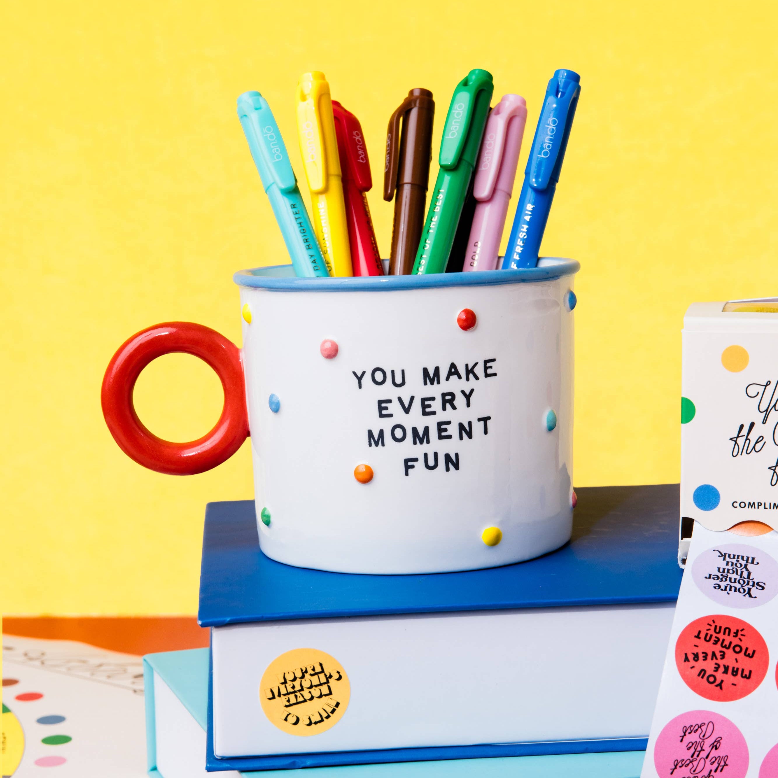 A white ceramic mug pen holder with a red handle, printed with the phrase 'YOU MAKE EVERY MOMENT FUN', filled with colorful pens. It is styled on top of stacked books with stickers against a bright yellow background.