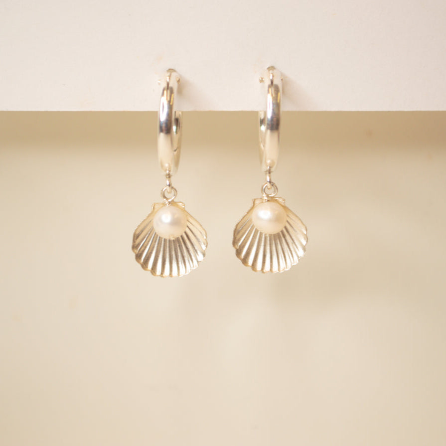Silver hoop with a scallop sea shell and pearl dangle on an off white background.