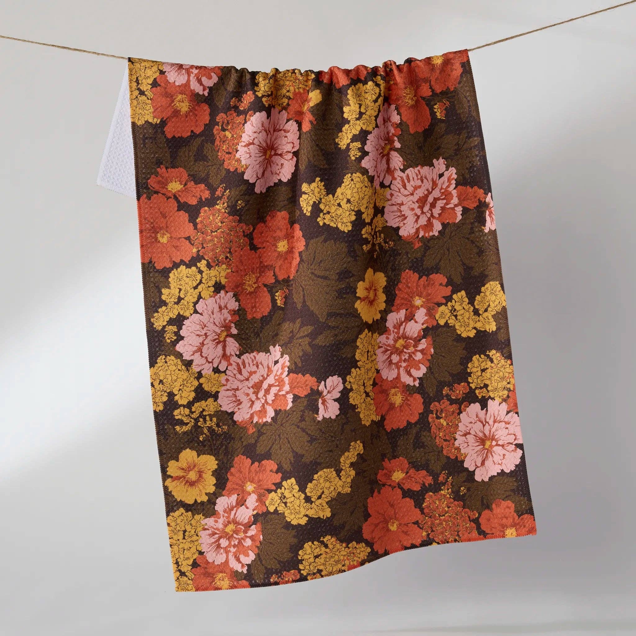 TEA TOWEL with a vintage floral pattern in orange, pink, and yellow on a dark brown background, hanging from a thin string against a light gray wall.