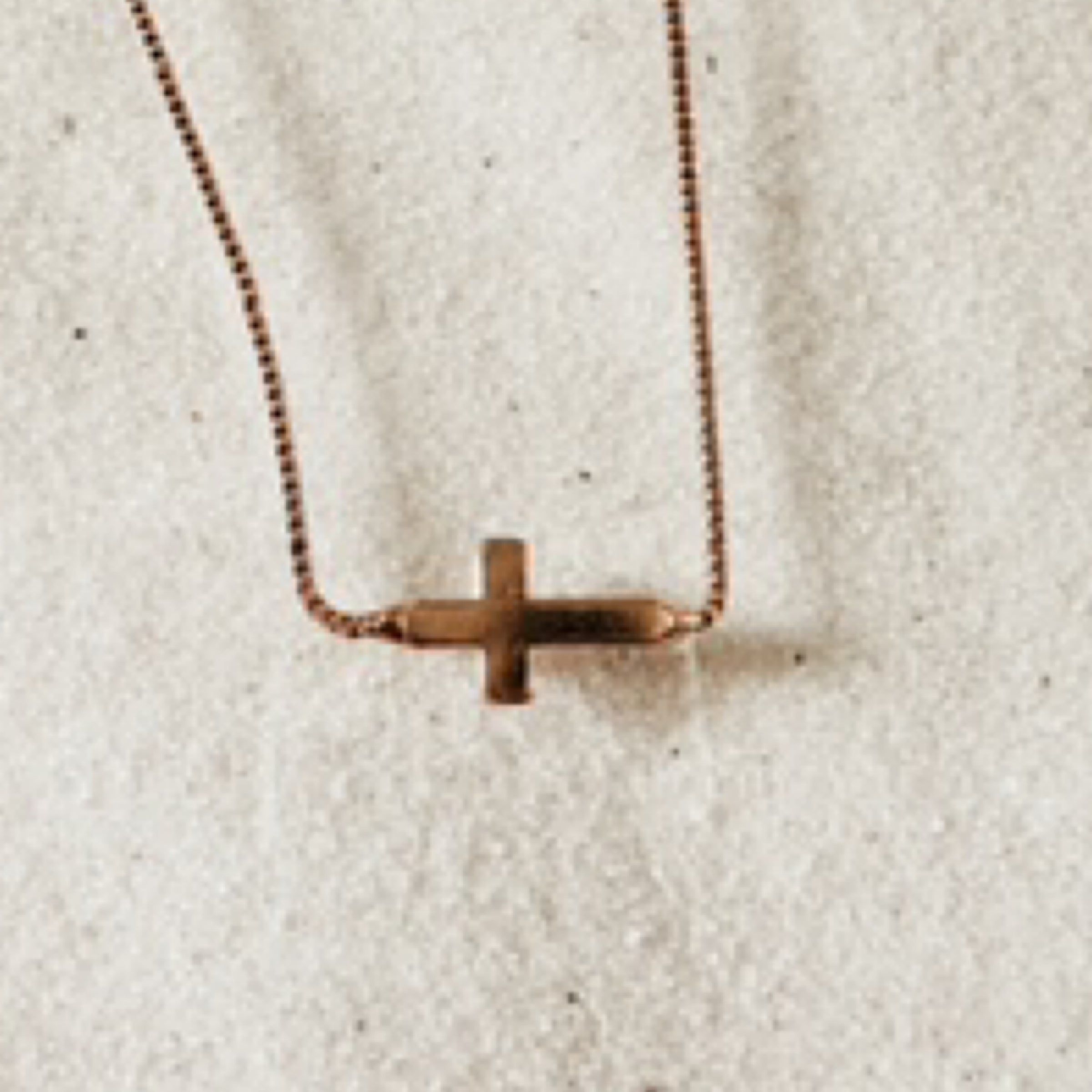 Simple gold chain with a gold cross hanging horizontally. Pictured against white sand.