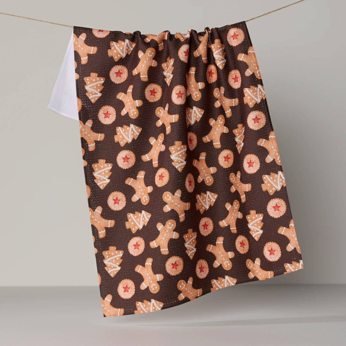 A dark brown kitchen towel with a gingerbread man and tree pattern, hanging from a string against a neutral gray background.