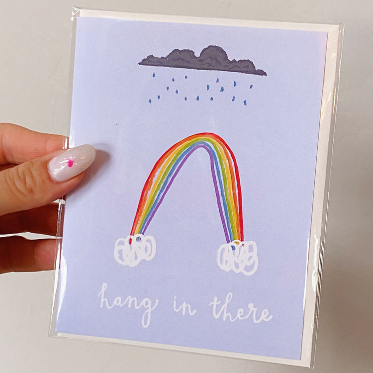 lavender colored card with a rainbow and rain cloud reading "hang in there"