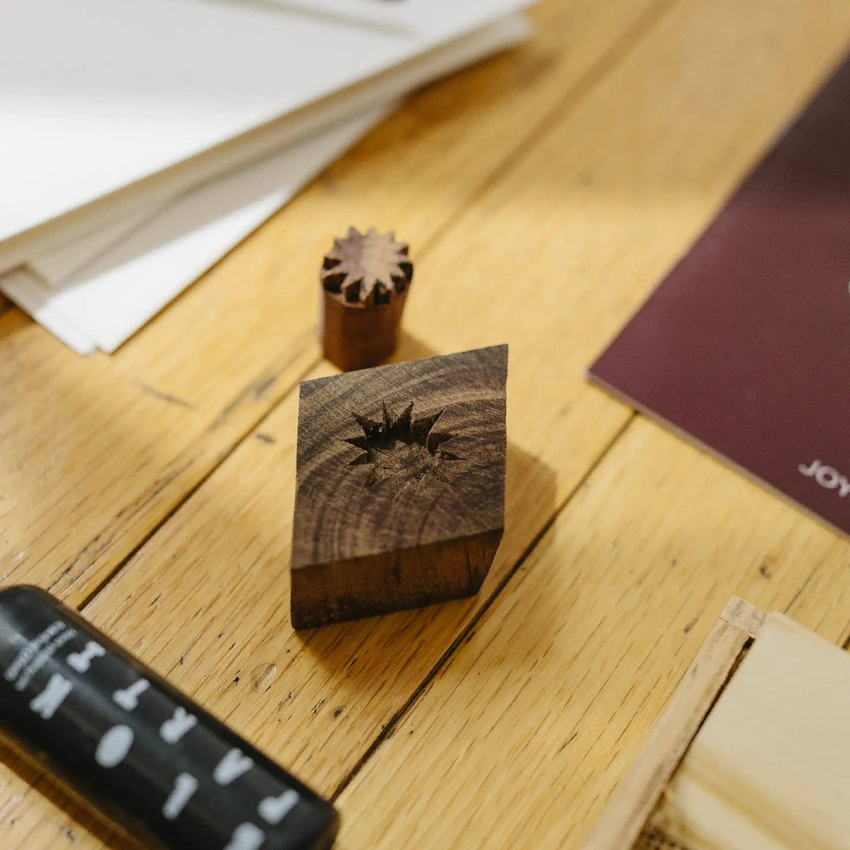 A wooden stamp block with a starburst design on top, shown on a light wood tabletop alongside other stationery items.