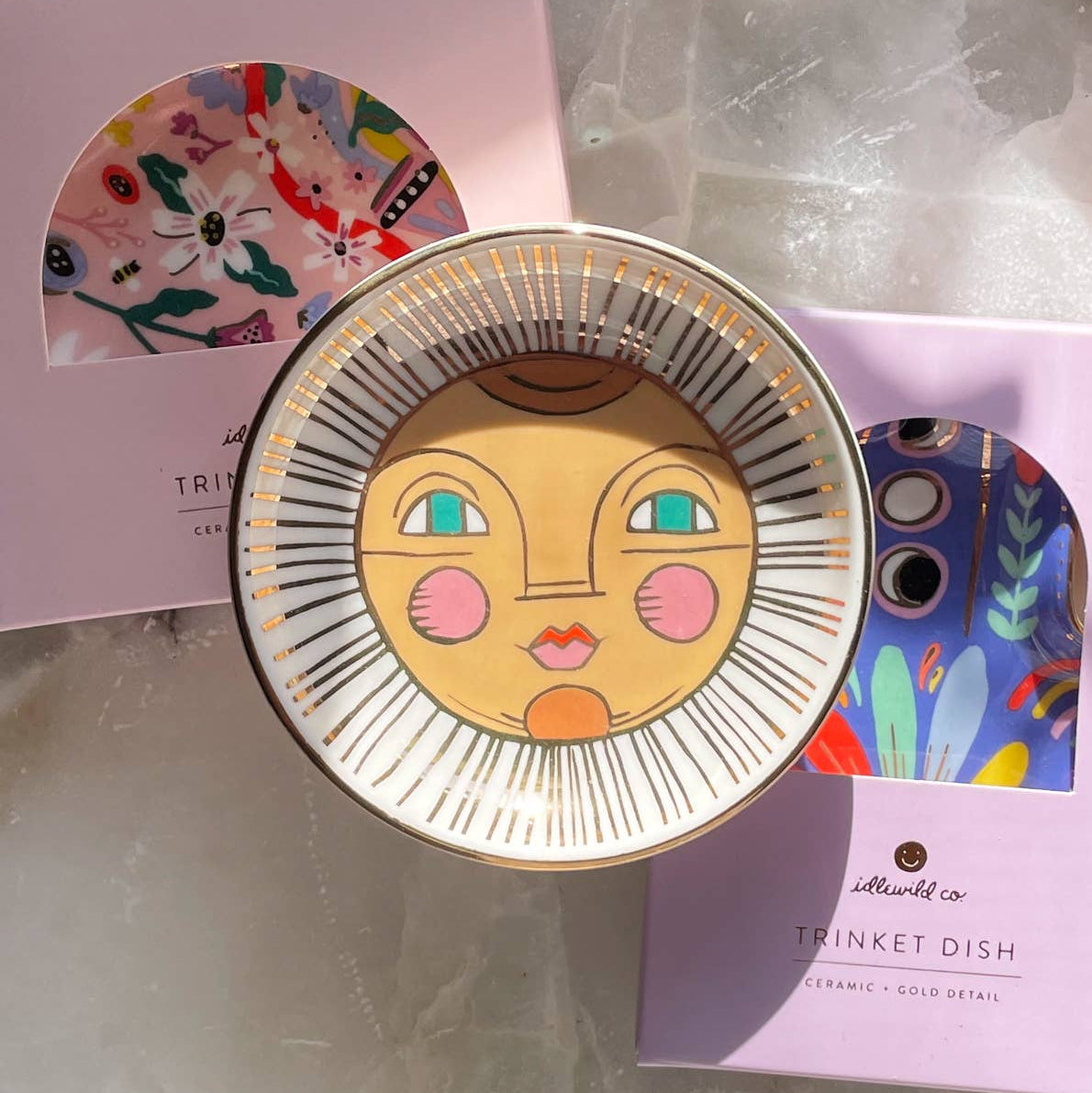 Decorative ceramic trinket dish with face design on a marble surface, with packaging in the background.