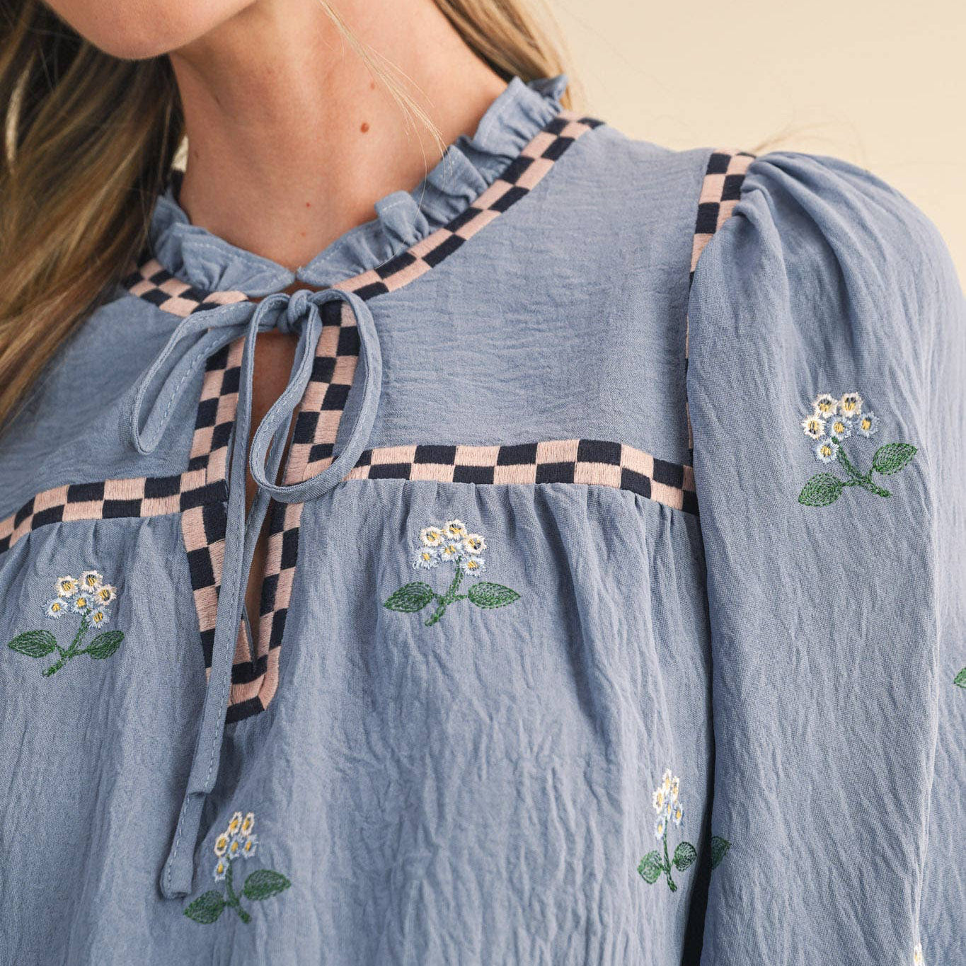 Embrace effortless charm with this enchanting floral embroidered babydoll top in soft dusty blue. The delicate all-over floral embroidery paired with checkered details on the neckline and yoke creates a romantic, thoughtfully designed piece that celebrates feminine style. The ruffled neckline features a sweet keyhole opening with tying detail, while long balloon sleeves with elastic cuffs offer a flattering, comfortable fit. Fully lined for confidence and ease.