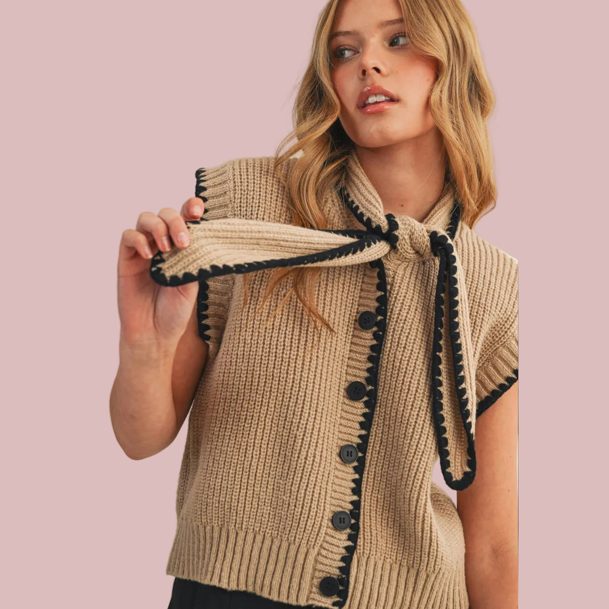 A woman wearing a beige ribbed knit cardigan vest with black trim and black buttons, holding up the matching tie-neck detail against a plain beige background.
