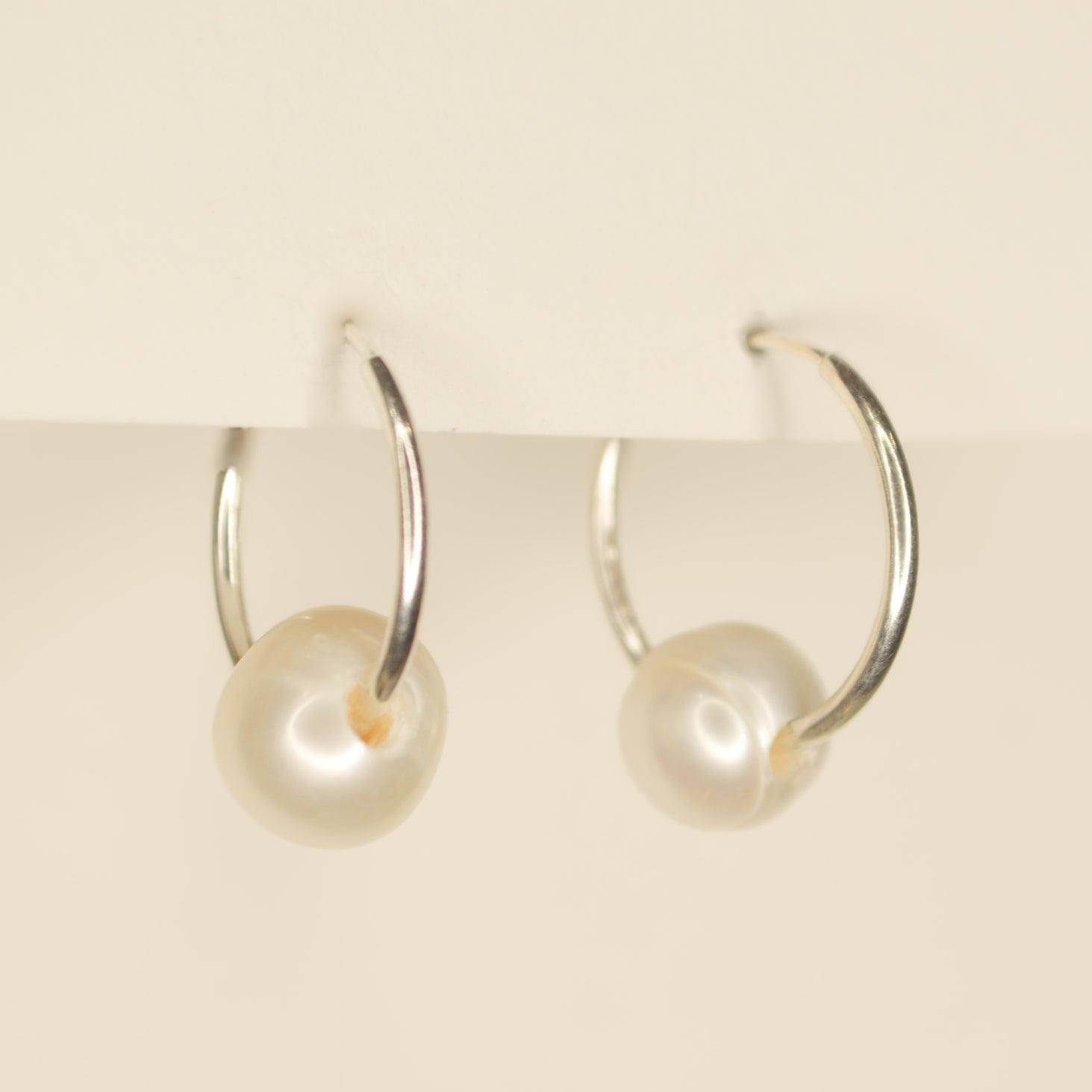 Thin silver hoops with a sliding white round pearl on each. Pictured on an off white background.