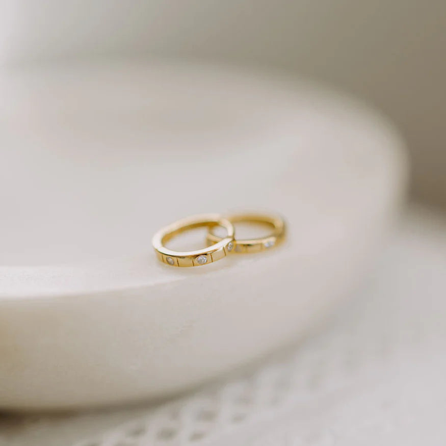 Two elegant gold rings with small embedded diamonds are displayed on a smooth, round cream-colored surface. The rings have a minimalist, modern design with evenly spaced stones, evoking a refined and timeless aesthetic.