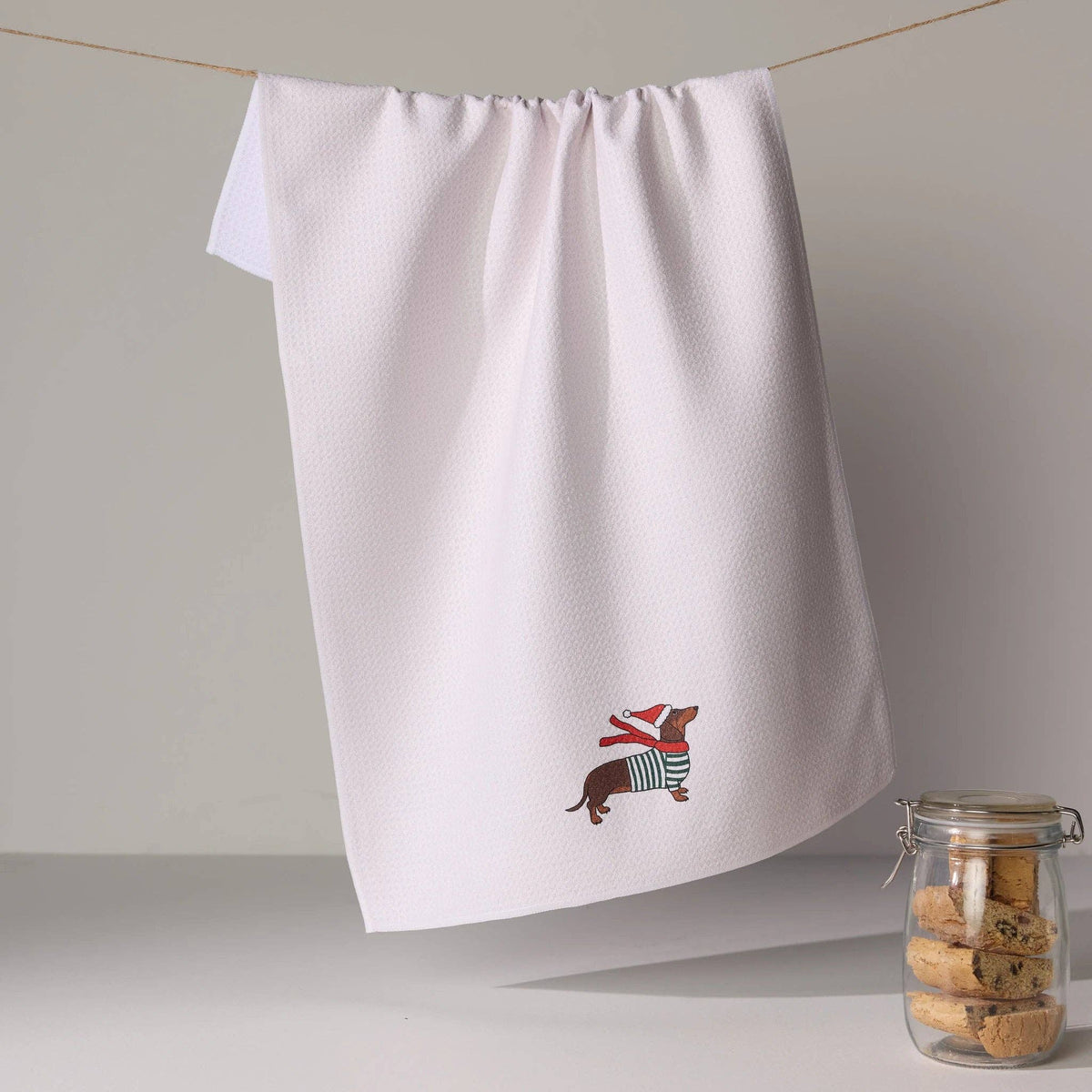 A white kitchen tea towel hangs from a string on a wall, featuring a small  dachshund wearing a red Santa hat and striped sweater near the bottom. A glass jar of cookies sits on the surface below.