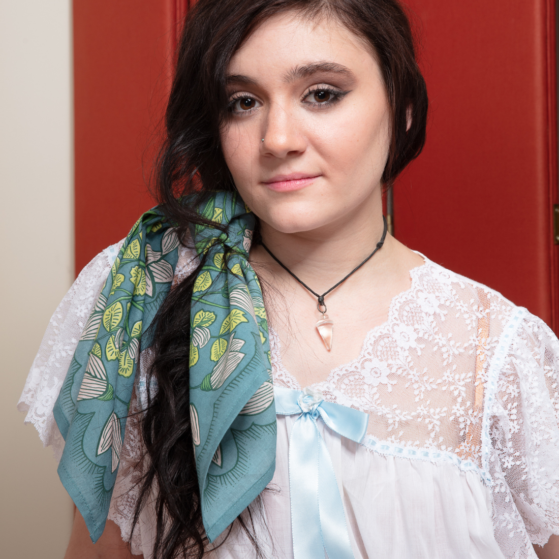 Woman wearing a white lace top and a blue floral scarf in front of a red door.
