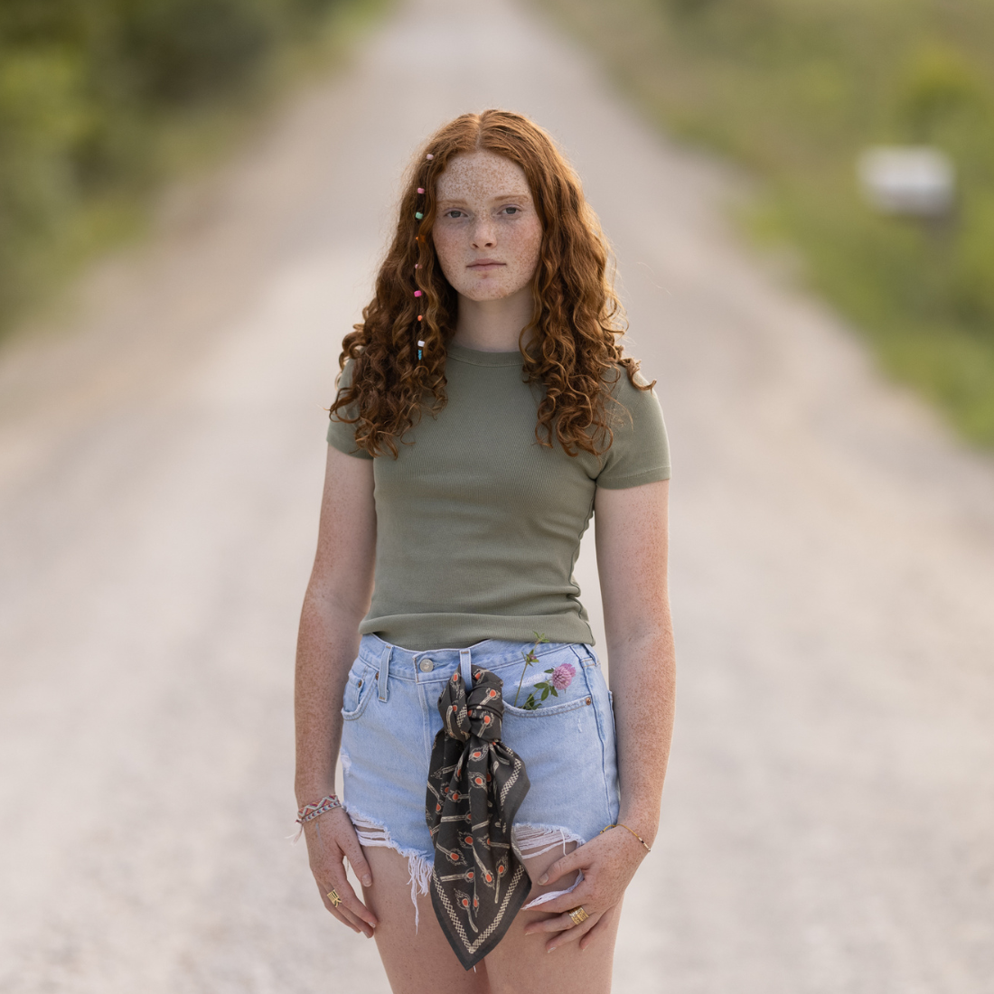Woman standing on a dirt road wearing a green shirt and denim shorts.