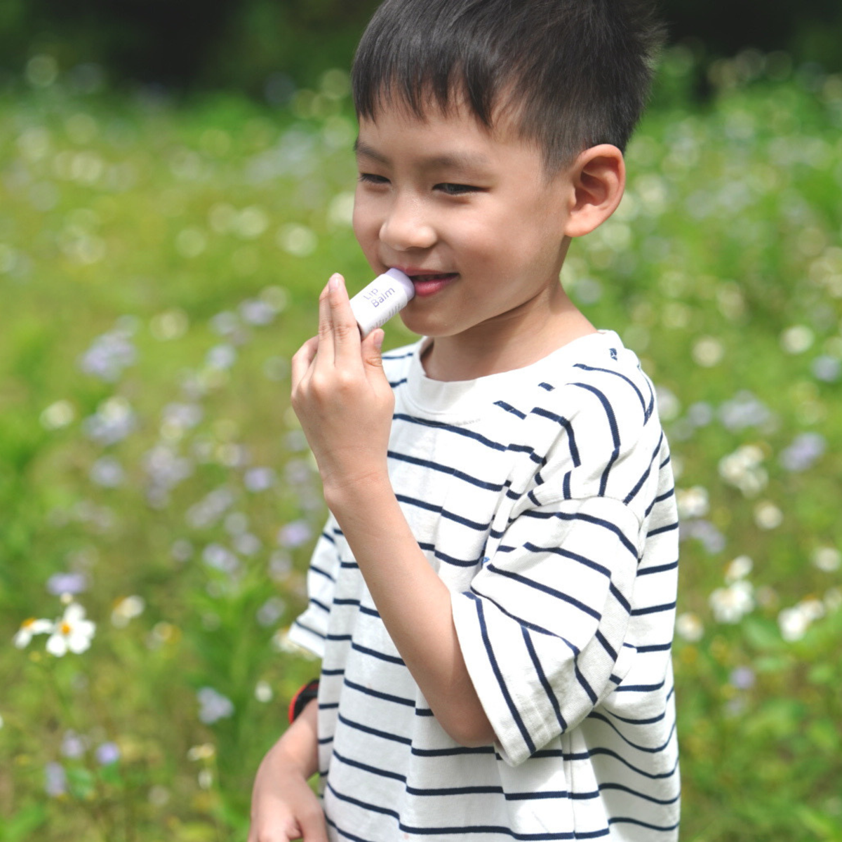 A young boy wearing a white t-shirt with navy blue horizontal stripes stands in a grassy field with small white flowers, holding a white cylindrical product to his mouth.