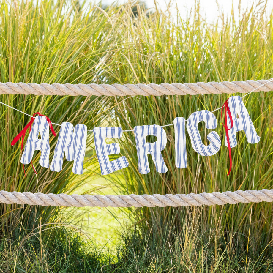 Celebrate in timeless style with the America Ticking Fabric Puffy Banner from our Hamptons collection, a charming addition to your Americana home and party décor. Crafted from soft linen and featuring puffy, blue-and-cream striped letters stitched together with cream twine and finished with bright red ribbon bows, this festive banner brings a cozy, patriotic touch to Fourth of July, Memorial Day, and all your summer gatherings.