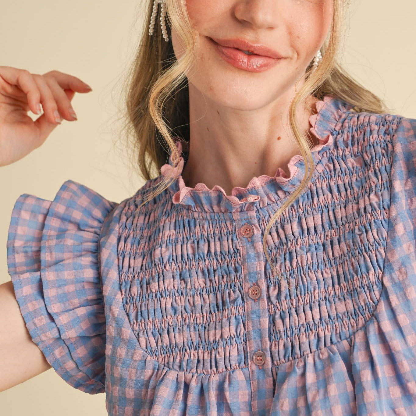 Embrace your softer side in this charming gingham flutter sleeve top. The delicate ruffled lace trim at the neckline and playful flutter sleeves create a romantic, feminine silhouette that feels both cheerful and flattering. A smocked yoke adds gentle structure, while the babydoll bodice offers comfortable, forgiving fit. The button-down placket makes it easy to style however you'd like—dress it up or keep it casual. Lined for confidence and comfort. Available in a lovely blue and pink.