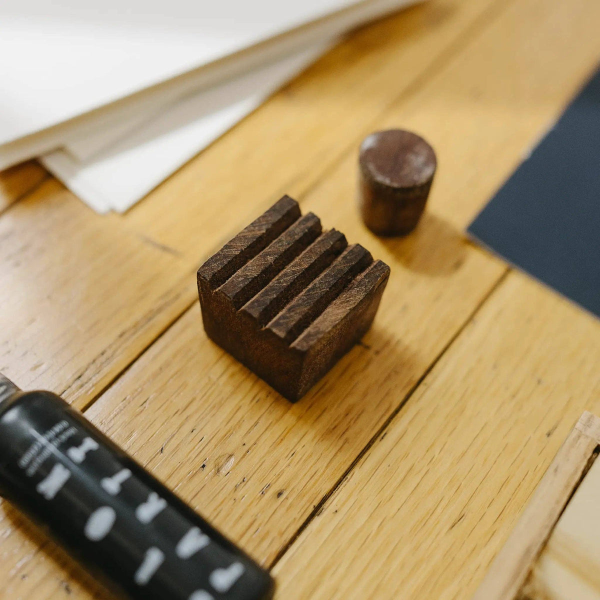 A dark brown wooden stamp block with a ridged top surface and a matching cylindrical wooden roller, displayed on a light wood tabletop alongside a black bottle and papers.