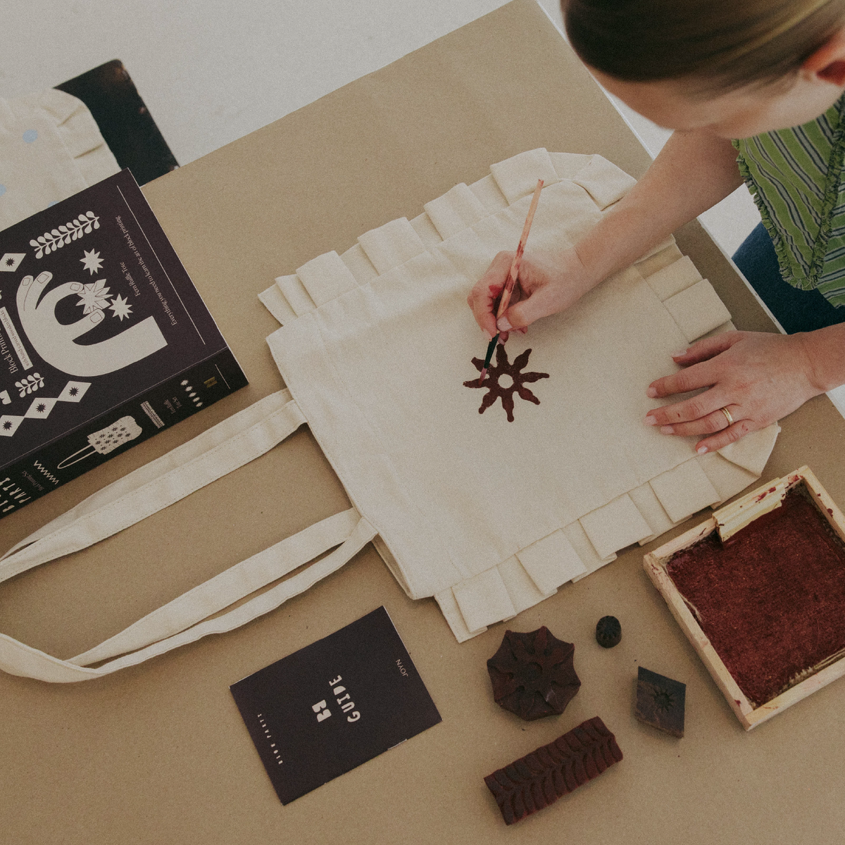 Person stamping a design on a tote bag with a wooden block and ink pad, surrounded by stationery items.