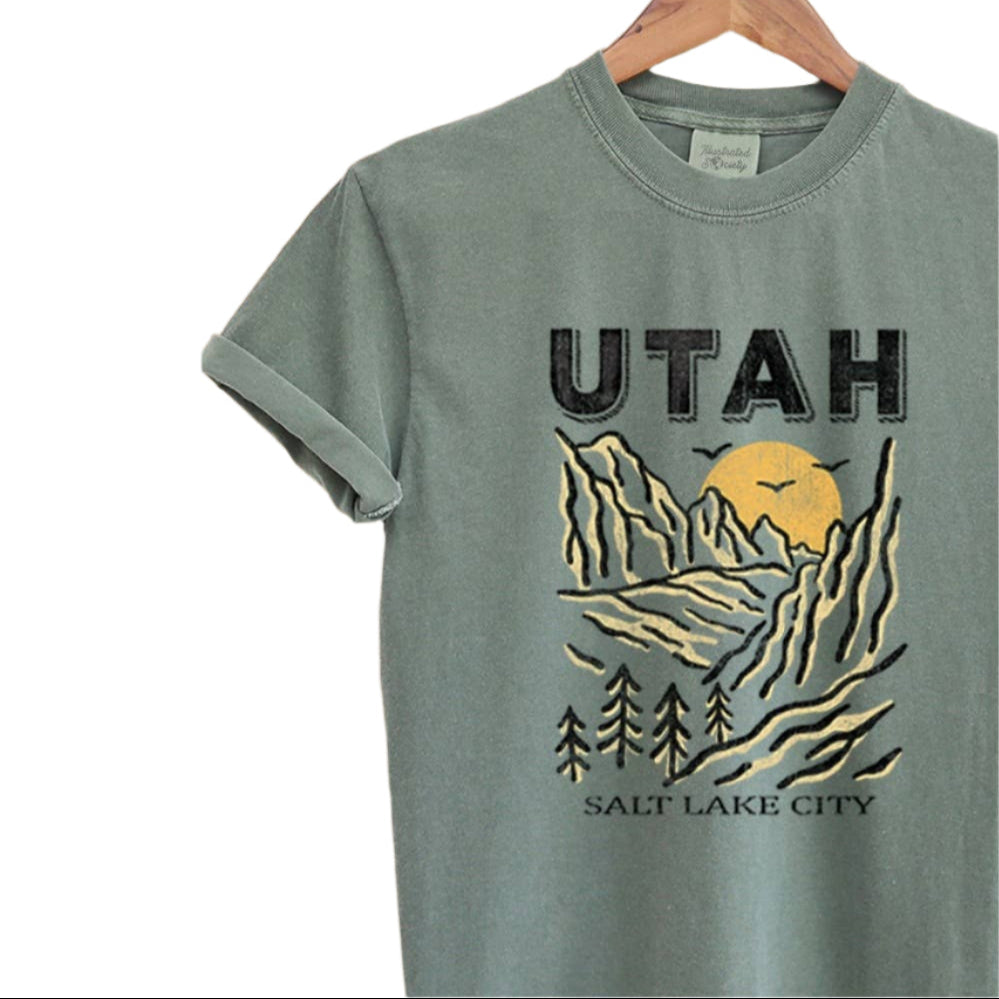 Green t-shirt with 'Utah' and mountain graphic on a hanger against a textured beige background.