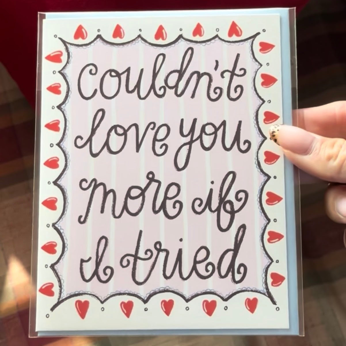 A heartfelt greeting card in a clear plastic sleeve displays the playful handwritten message "Couldn't love you more if I tried" in elegant black cursive lettering, centered on a light pink background with subtle vertical stripes.

The card is bordered by a scalloped frame adorned with numerous small red hearts and tiny white dots, creating a sweet, romantic, and affectionate design perfect for expressing deep love or appreciation.