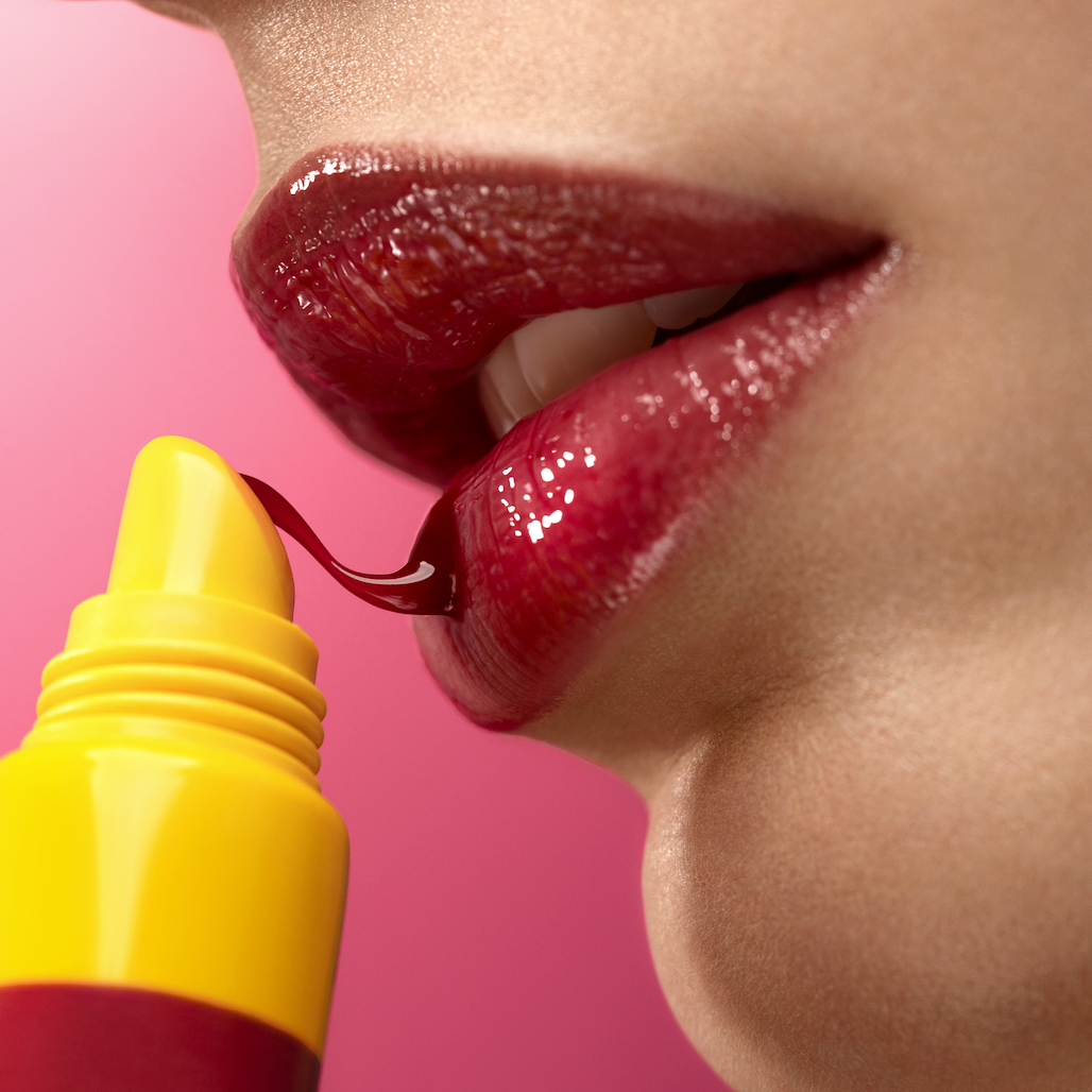 Close-up of a person applying red lipstick with a yellow applicator on a pink background