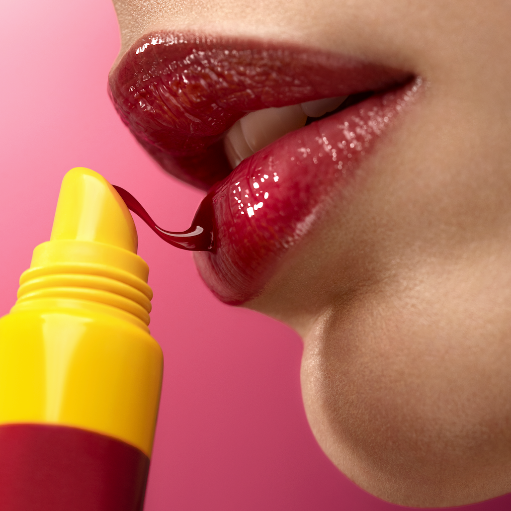 Close-up of a person applying red lipstick with a yellow applicator on a pink background