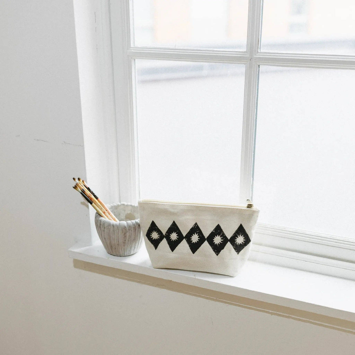 A cream canvas pouch with black diamond and star patterns sits on a white windowsill next to a small gray cup holding pencils.