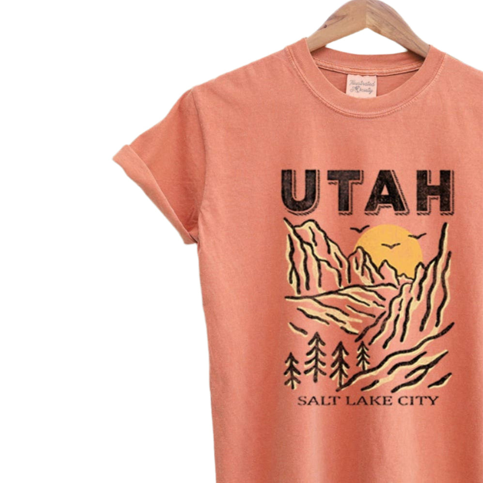 Coral t-shirt with 'Utah' and 'Salt Lake City' graphic on a hanger against a textured beige background.