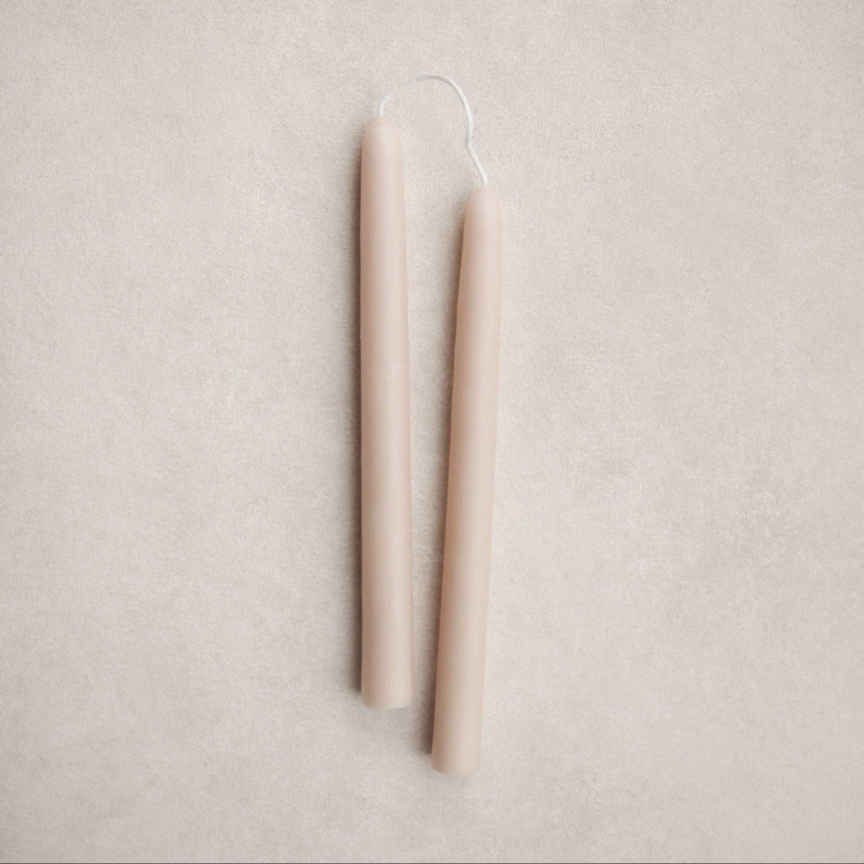 Two slim, tapered candles in a warm salt tone, shown upright on a textured light beige background.