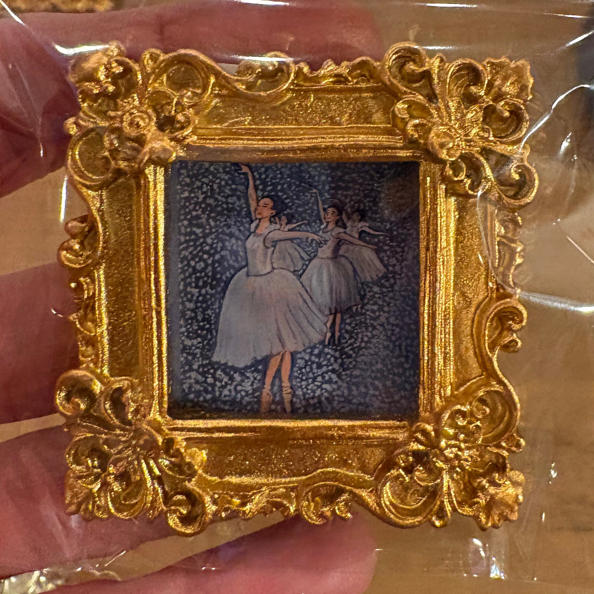 Gold decorative frame with a miniature painting inside, held by a hand against a neutral background.
